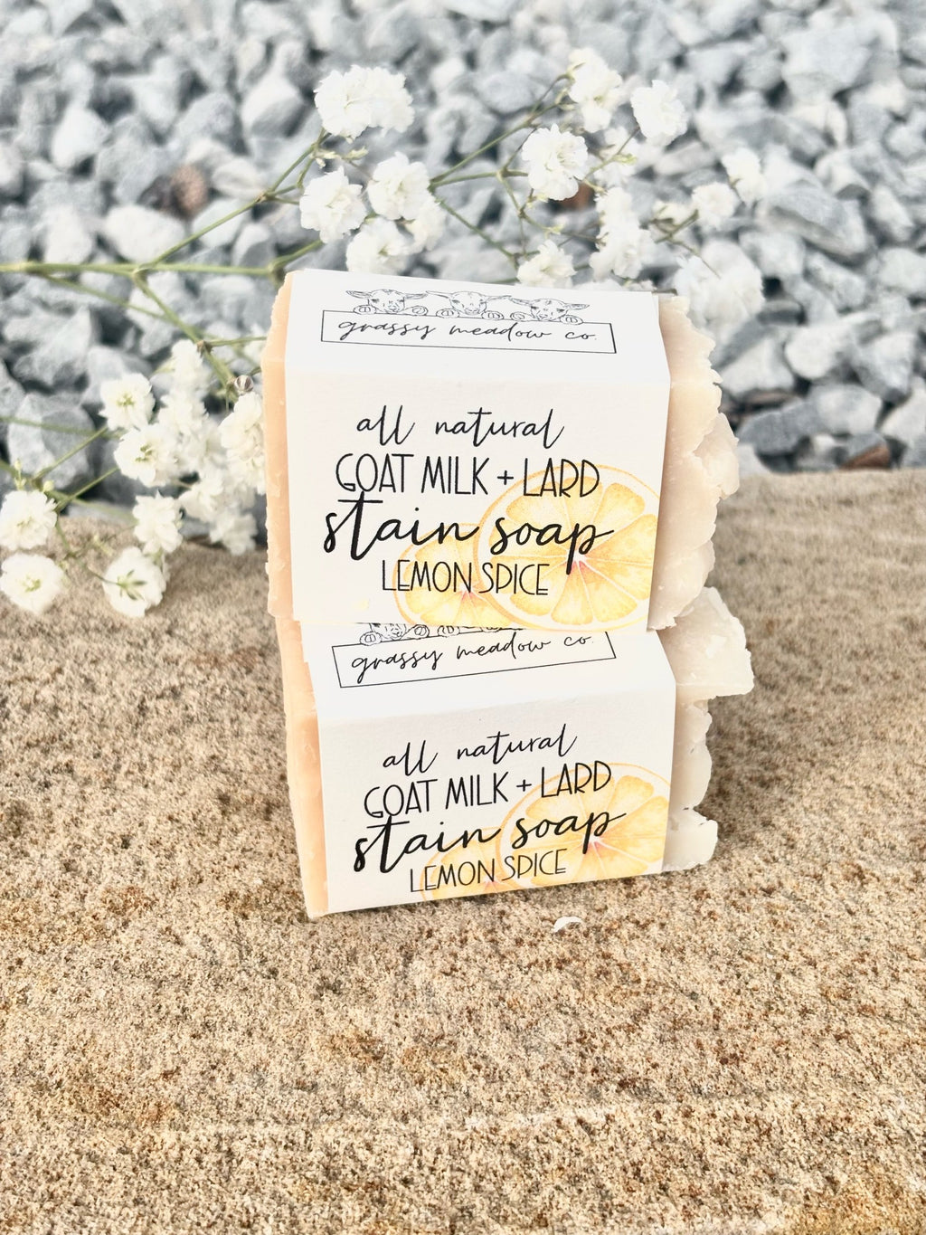 Two bars of lemon spice stain soap stacked on a sandy surface with white flowers and gray pebbles in the background. The soap bars are wrapped in white paper labels with black text and a yellow lemon slice graphic.