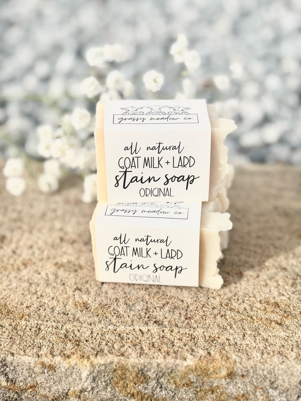 Two handmade soap bars wrapped in white paper labels reading 'all natural GOAT MILK + LARD stain soap Original' by grassy meadow co, displayed on a sandy surface with a blurred background of white flowers.