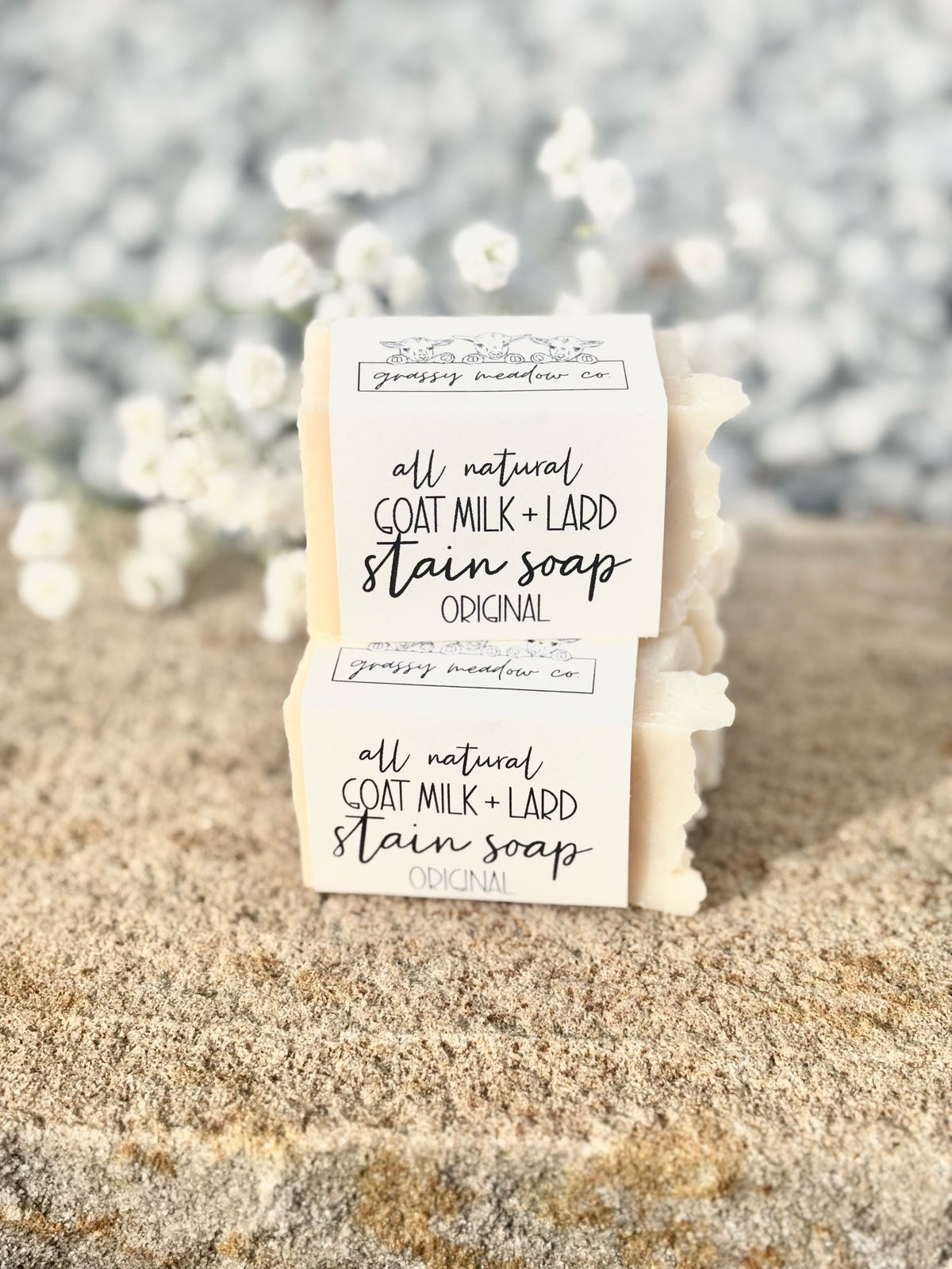 Two handmade soap bars wrapped in white paper labels reading 'all natural GOAT MILK + LARD stain soap Original' by grassy meadow co, displayed on a sandy surface with a blurred background of white flowers.