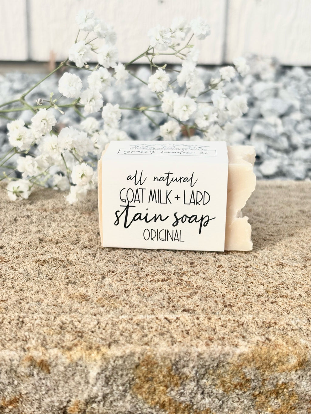 A bar of goat milk and lard stain soap in the 'Original' scent, wrapped in a white paper sleeve with black text, resting on a textured stone surface with white baby's breath flowers in the background.