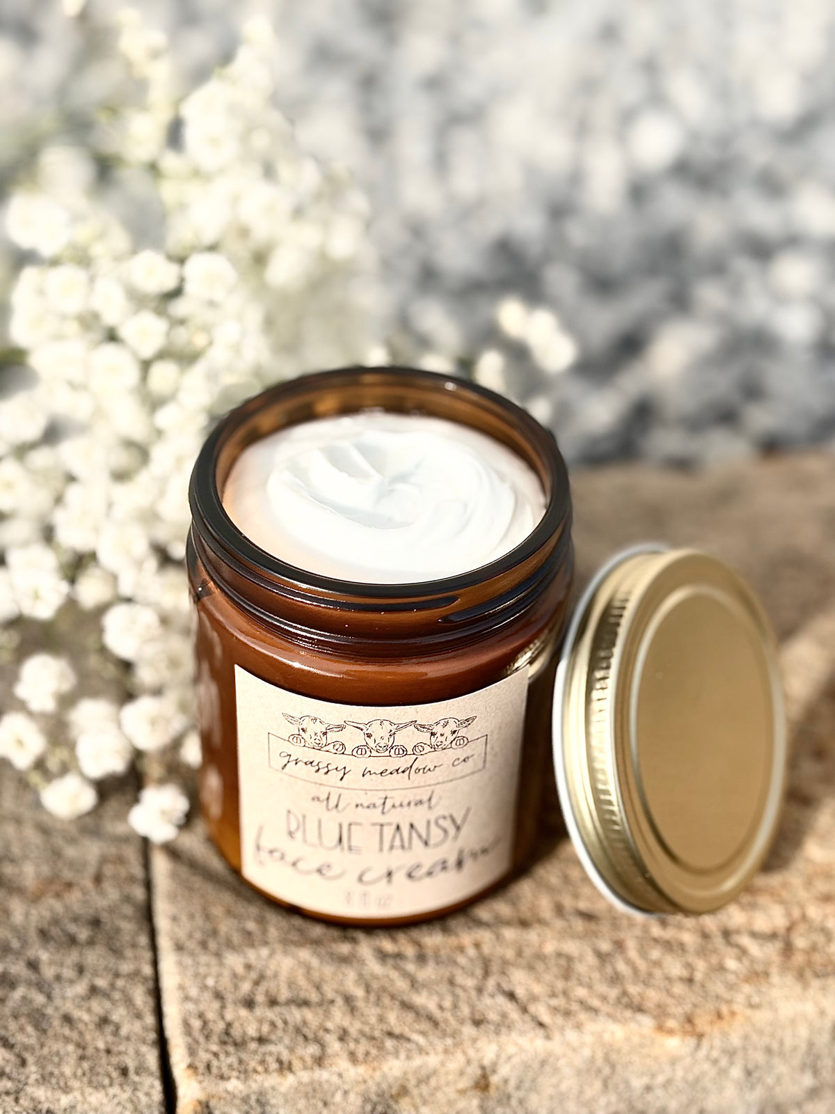 An open amber glass jar of Grassy Meadow All Natural Blue Tansy Face Cream, showing the white cream inside, with its gold lid resting beside it on a rustic wooden surface.