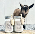 Two mason jar laundry detergents labeled 'all-natural' with small wooden scoops tied to the jars, set on a stone surface in front of a small goat standing against a white paneled wall.