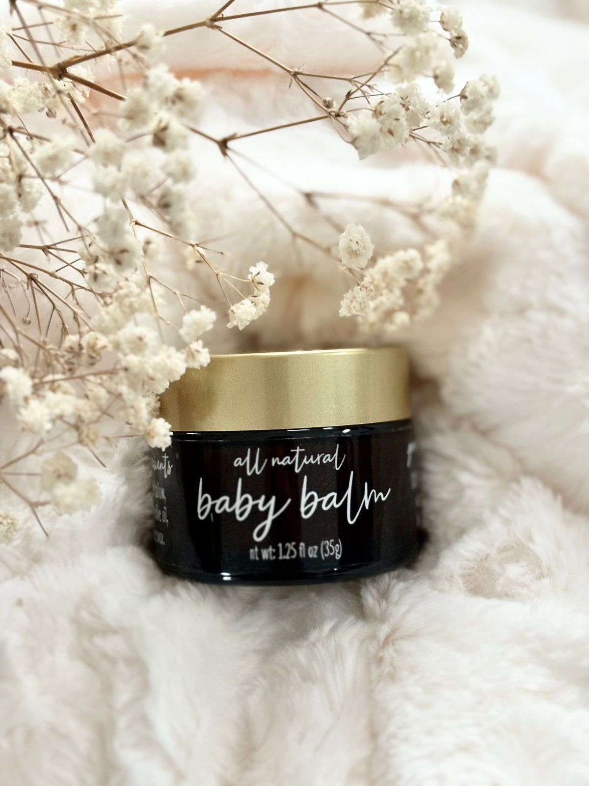 A small black jar of all natural baby balm with a metallic gold lid, styled on a soft white faux fur surface with dried white baby's breath flowers.