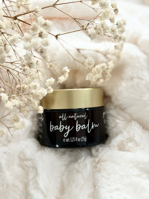 A small black jar of all natural baby balm with a metallic gold lid, styled on a soft white faux fur surface with dried white baby's breath flowers.