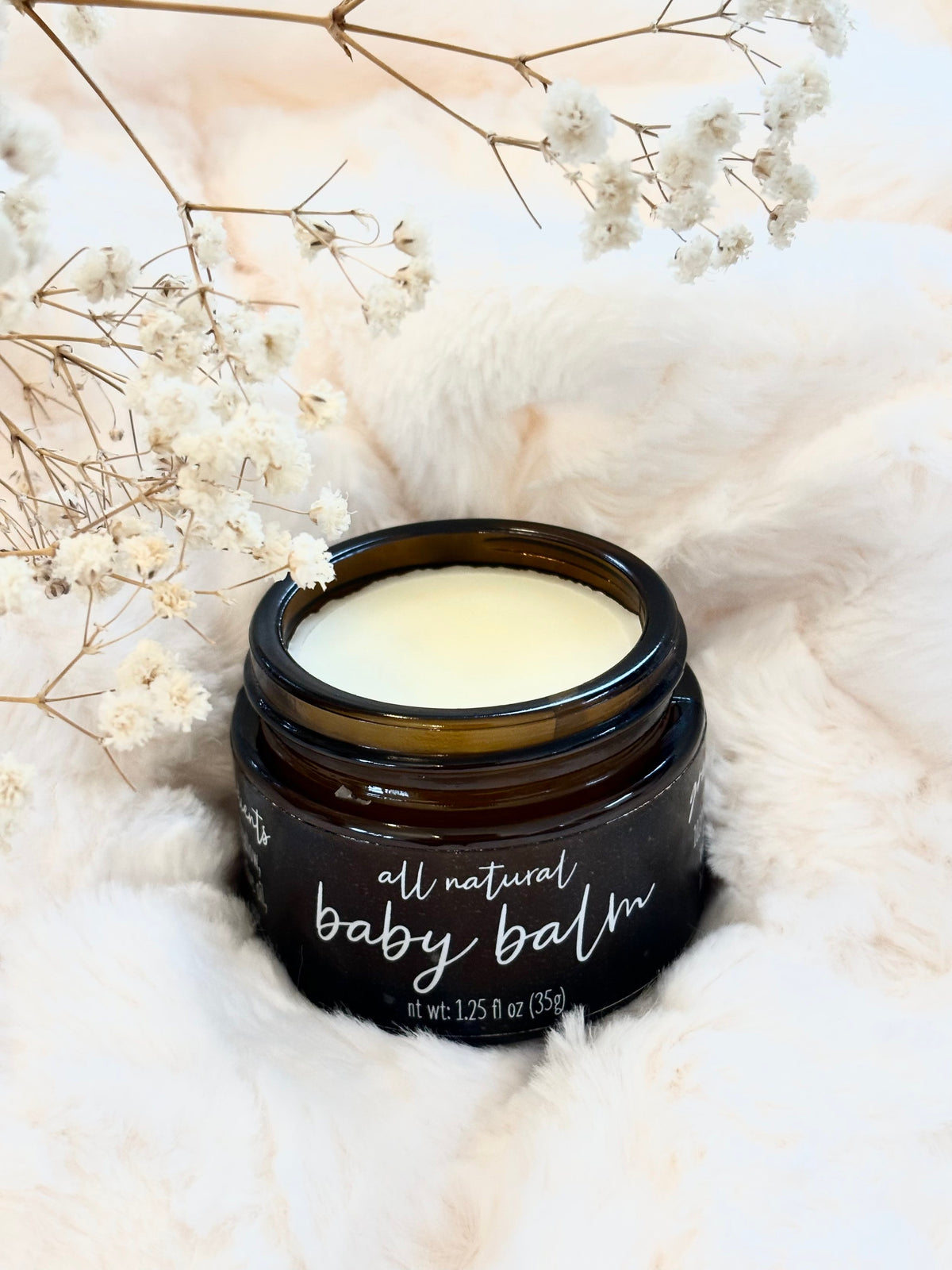 An open dark amber glass jar of baby balm with white cream inside, labeled 'all natural baby balm,' styled on a soft white faux-fur surface with dried white flowers around it.