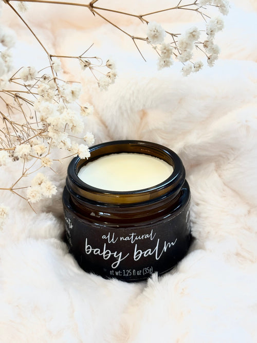 An open dark amber glass jar of baby balm with white cream inside, labeled 'all natural baby balm,' styled on a soft white faux-fur surface with dried white flowers around it.