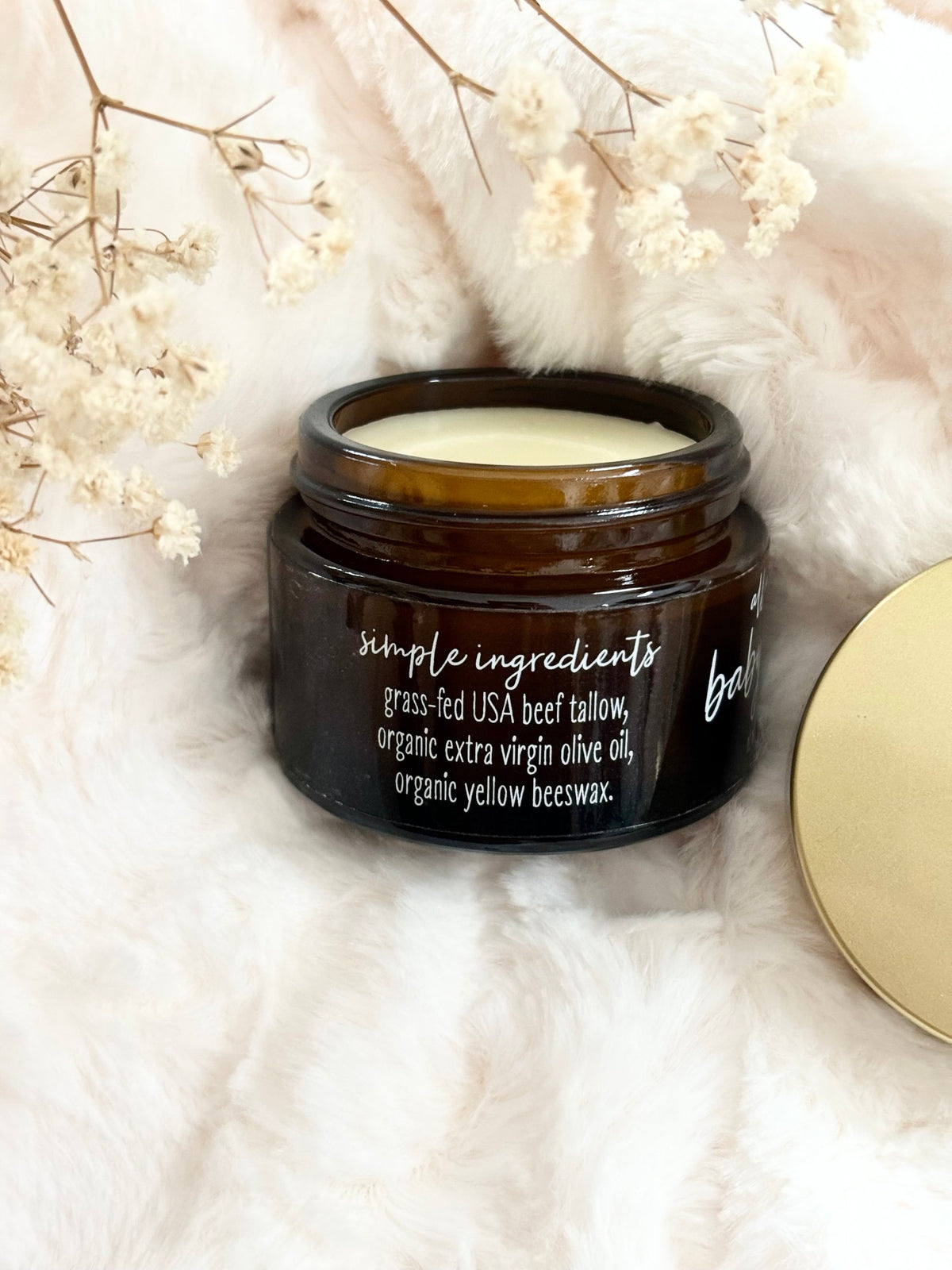 An amber glass jar of Simple Ingredients Beeswax Baby Balm with its gold lid resting beside it on a white faux fur surface, decorated with dried white flowers.