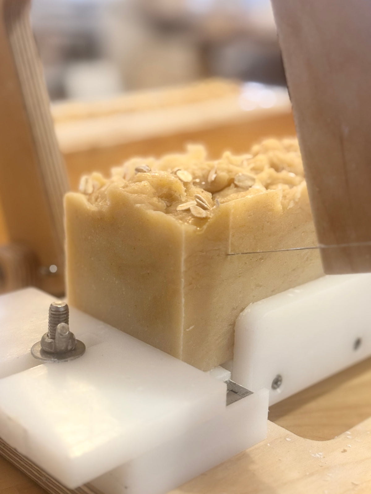 A close-up of a handmade oatmeal-colored soap bar with rolled oats on top, being sliced by a wooden and metal artisanal soap cutter.