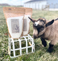 Two stand-up pouches of all-natural Laundry Detergent by Grassy Meadow Co. are displayed on a small white wire crate in a grassy field, with a small goat standing beside them.