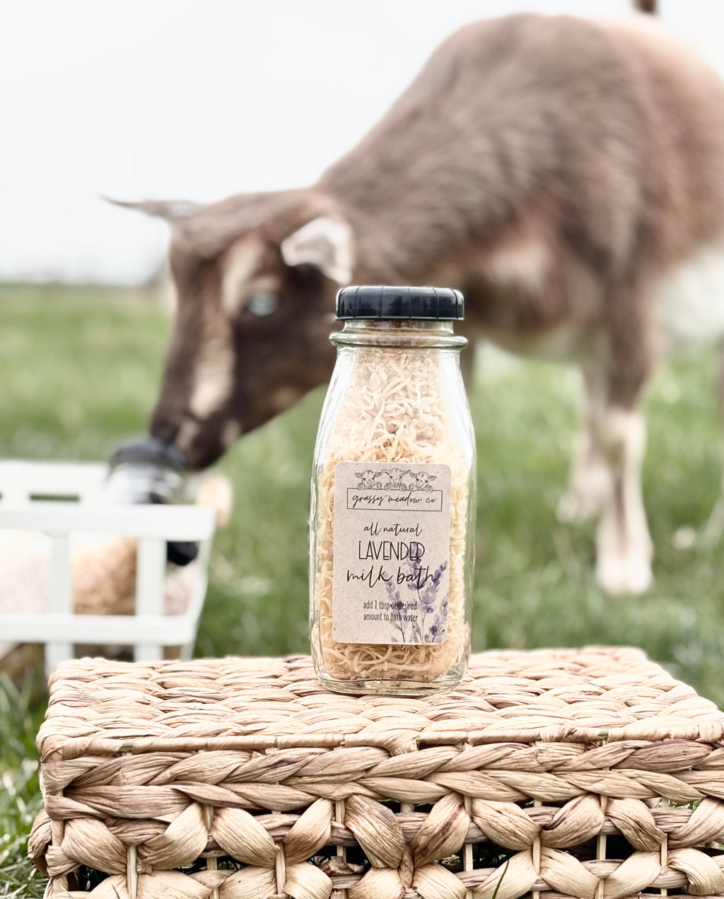 A glass bottle of Lavender Bubble Bath with dried lavender buds, set on a woven wicker basket outdoors with a goat and grassy field in the background.