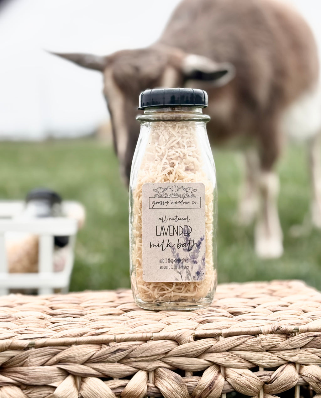 A glass bottle of lavender milk bath sits on a woven wicker surface with a blurred goat in a grassy field in the background.