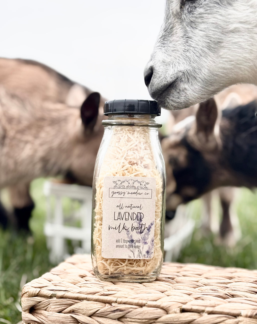 A glass jar of lavender milk bath sits on a woven basket with goats in a grassy outdoor setting behind it. The goats are out of focus, and the product jar is in sharp foreground detail.
