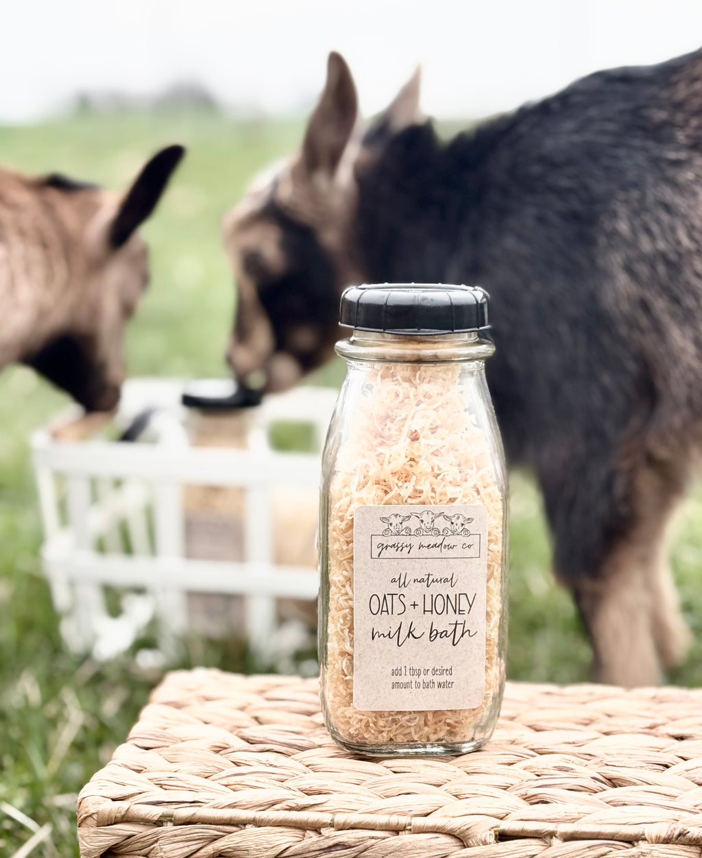 A glass bottle of grassy meadow co. all natural oats + honey milk bath sits on a woven straw mat, with two goats grazing in a blurred green field in the background.