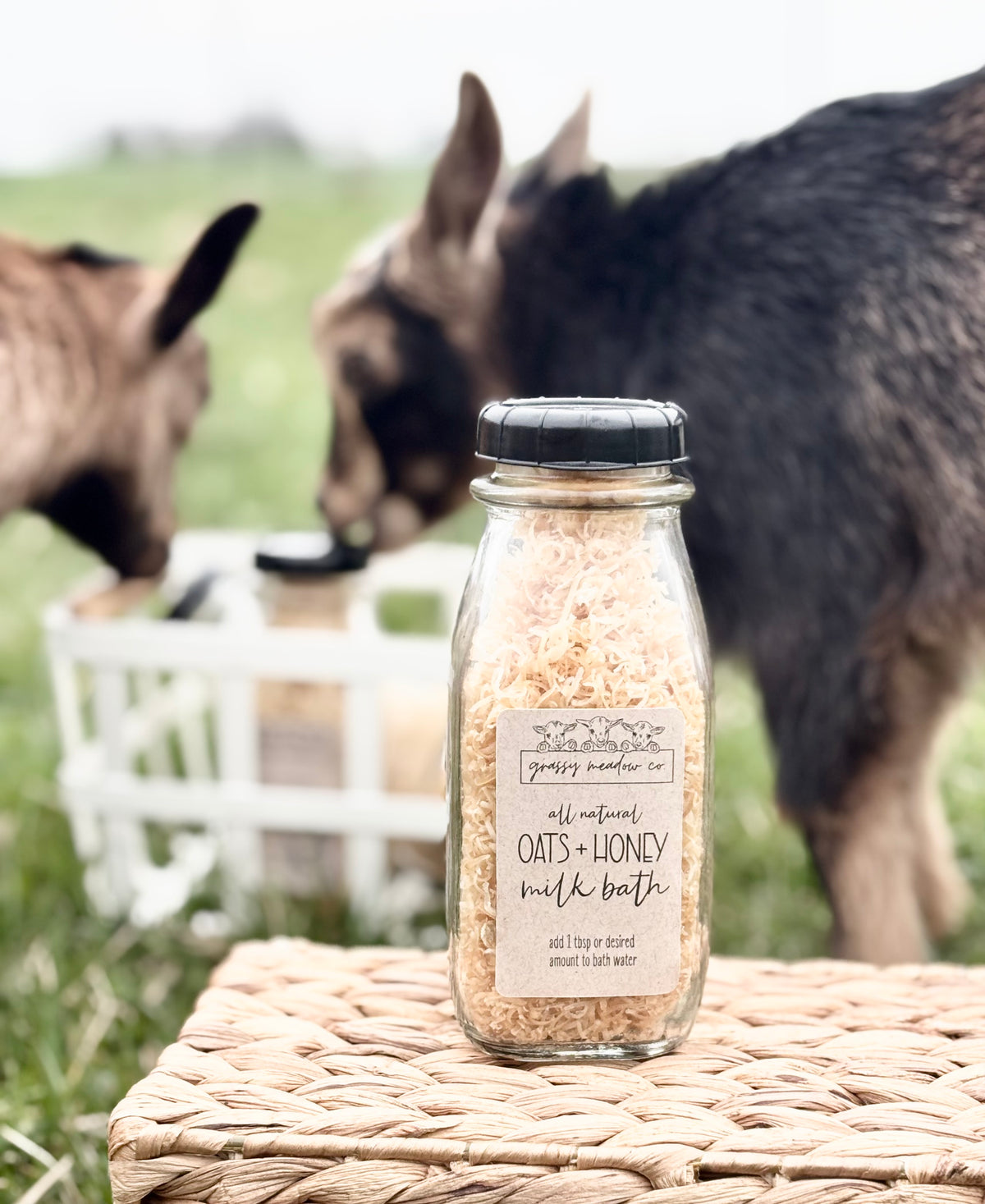 A glass bottle of grassy meadow co. all natural oats + honey milk bath sits on a woven straw mat, with two goats grazing in a blurred green field in the background.