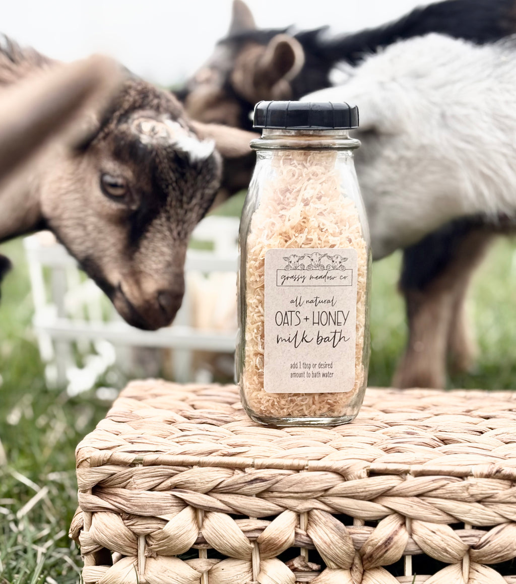 A glass jar of Grassy Meadow Co. Oats + Honey Milk Bath sits on a woven wicker basket outdoors, with two goats blurred in the background.