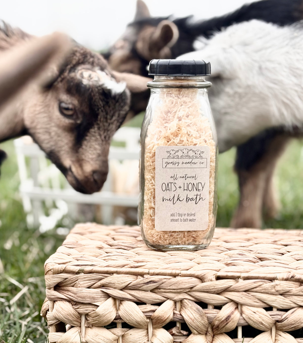 A glass jar of Grassy Meadow Co. Oats + Honey Milk Bath sits on a woven wicker basket outdoors, with two goats blurred in the background.