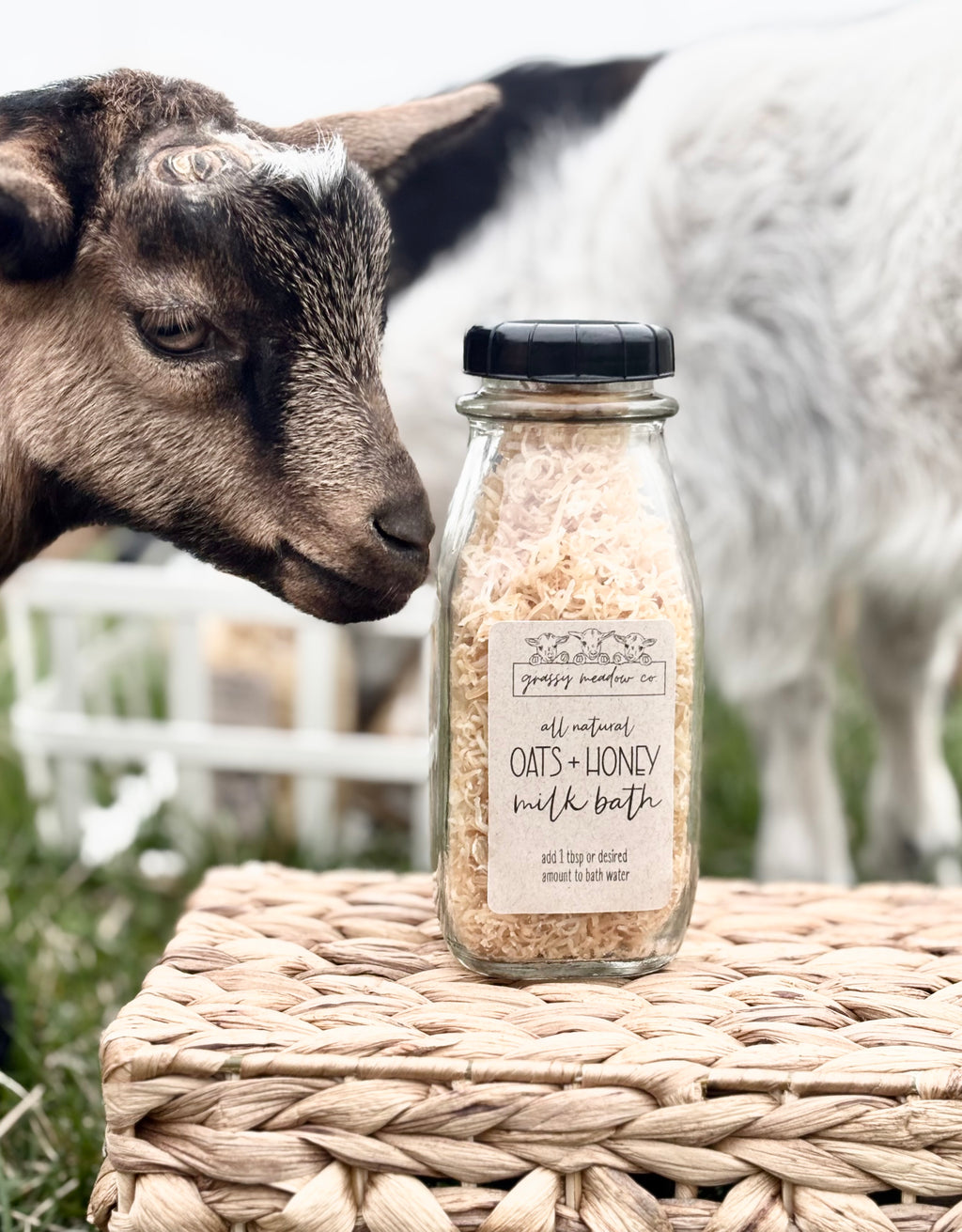 A clear glass bottle of Grassy Meadow Co. All Natural Oats + Honey Milk Bath sits on a woven wicker basket outdoors, with a goat head on the left and a silver goat in the background.