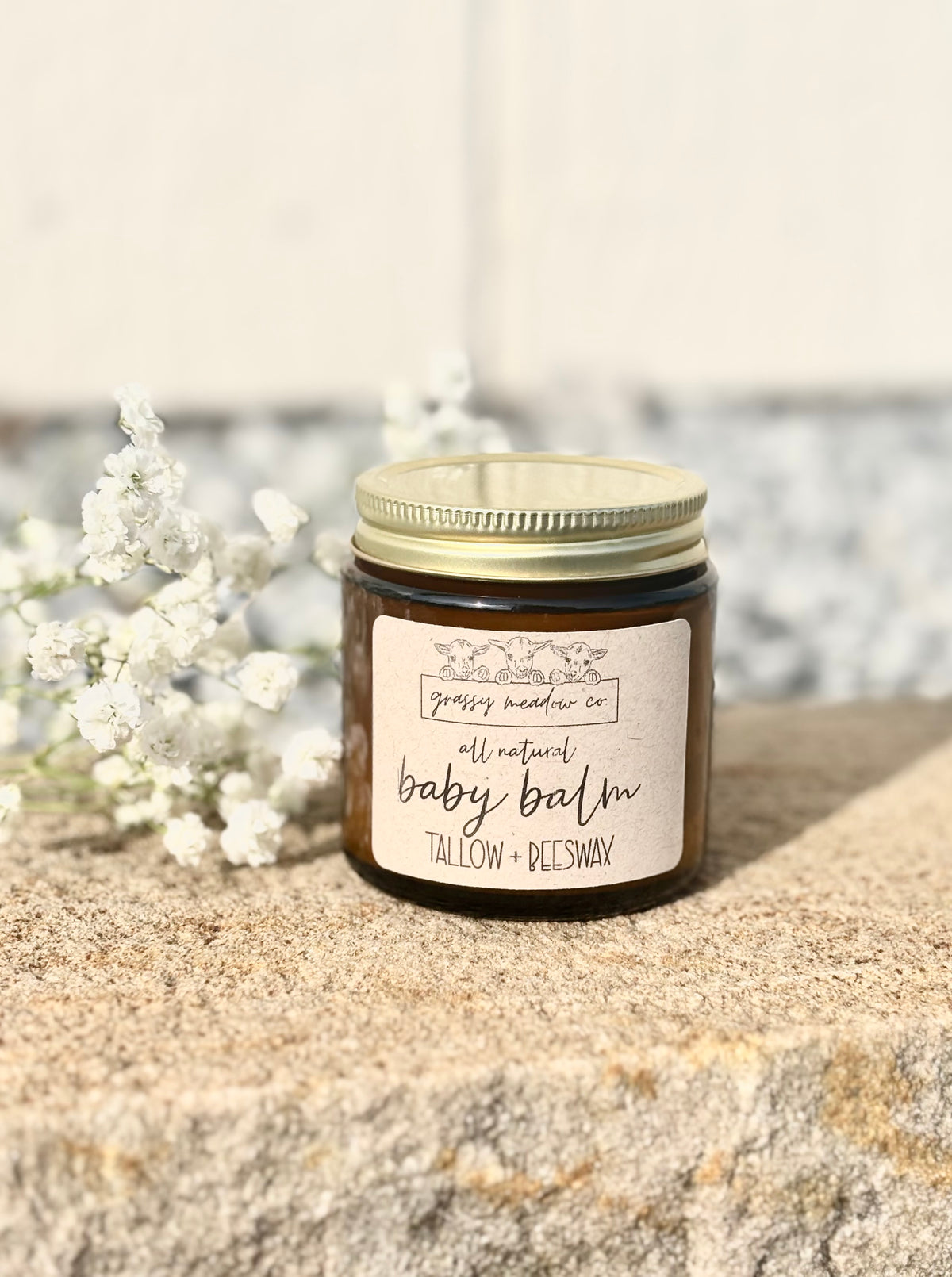 A small amber glass jar of baby balm with a gold metal lid and a cream label reading 'Grassy Meadow Co all natural baby balm TALLOW + BEESWAX,' placed on a stone surface with white flowers in the background.