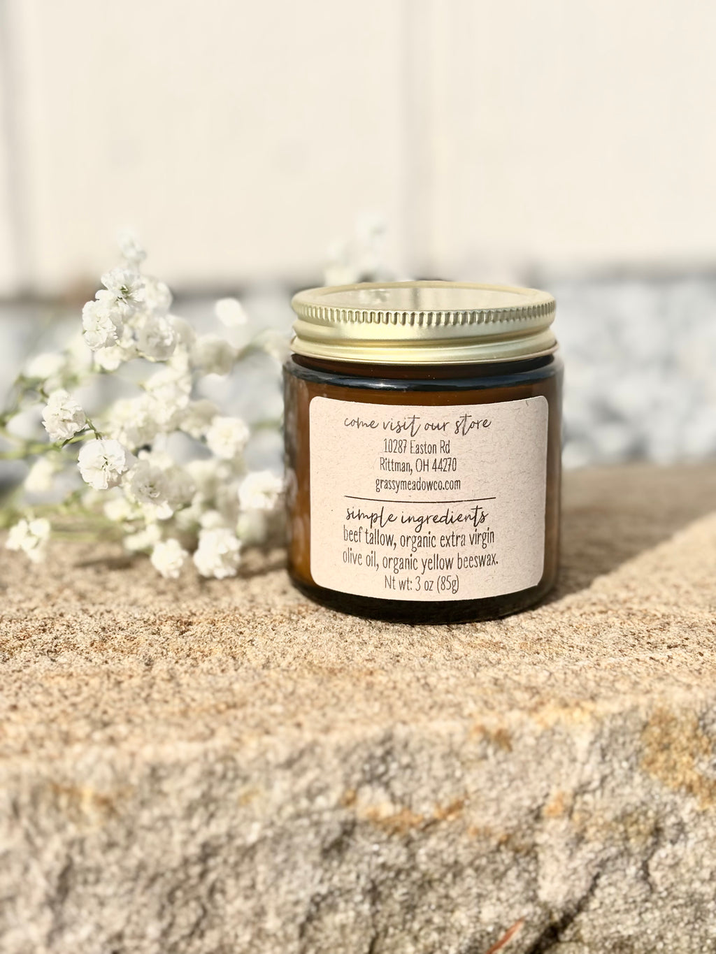 A small amber glass jar with a gold metal lid and a cream paper label, placed on a textured stone surface with white flowers in the background.