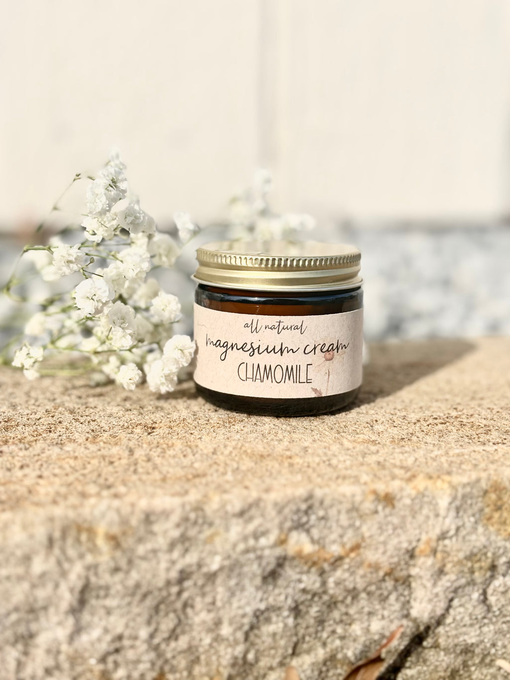A small glass jar of chamomile magnesium cream with a gold lid and a cream-colored label, styled on a textured stone surface next to white baby's breath flowers.