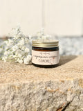 A small glass jar of chamomile magnesium cream with a gold lid and a cream-colored label, styled on a textured stone surface next to white baby's breath flowers.