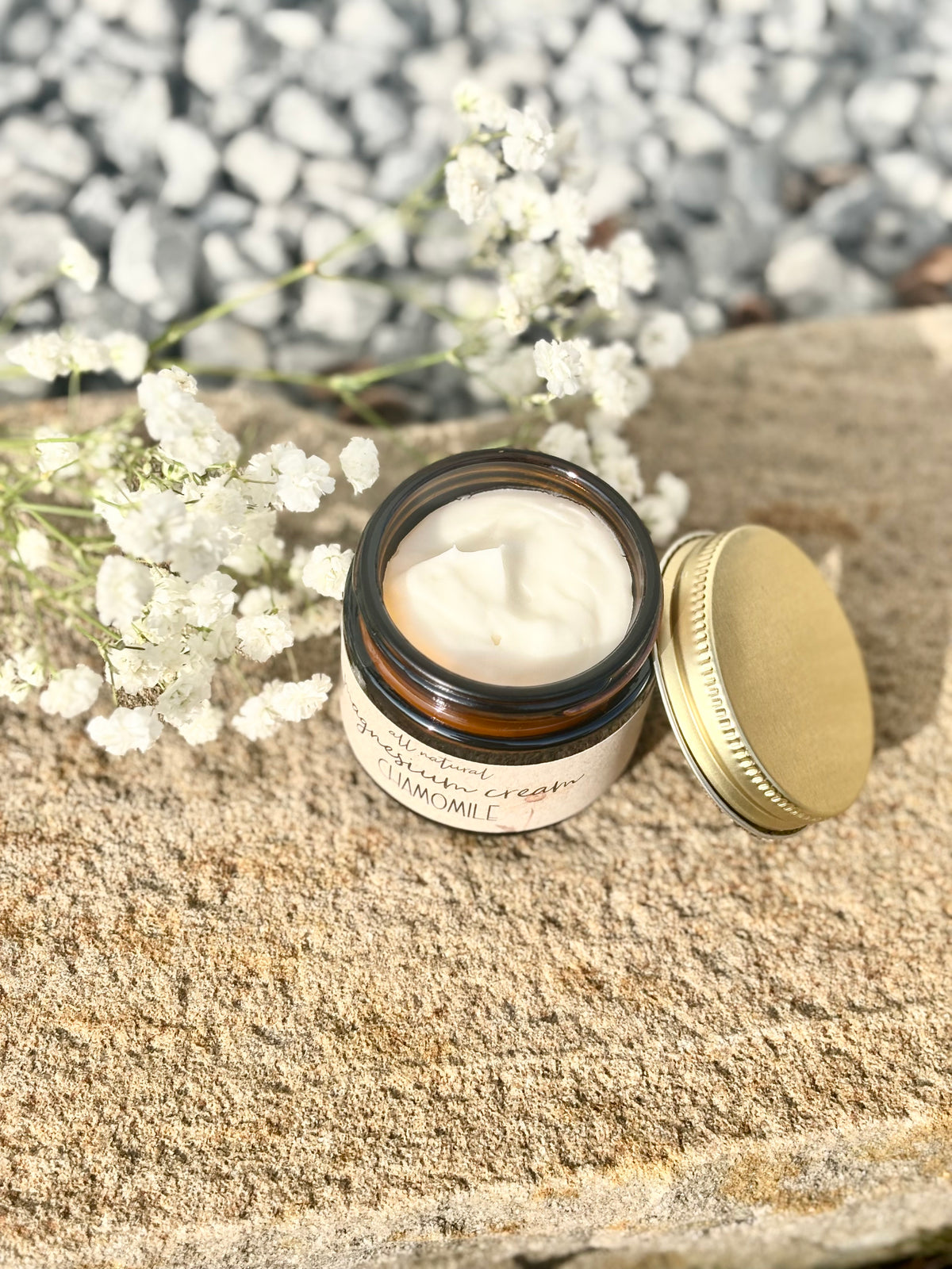 An open amber glass jar of body cream with a gold lid resting beside it on a stone surface, with white baby's breath flowers and a blurred pebble background.