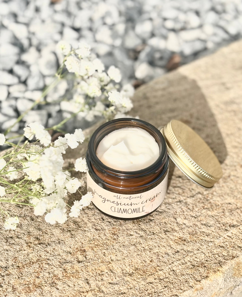 A small amber glass jar of claymole cream with its gold lid resting beside it on a stone surface, surrounded by white baby's breath flowers and a blurred gravel background.