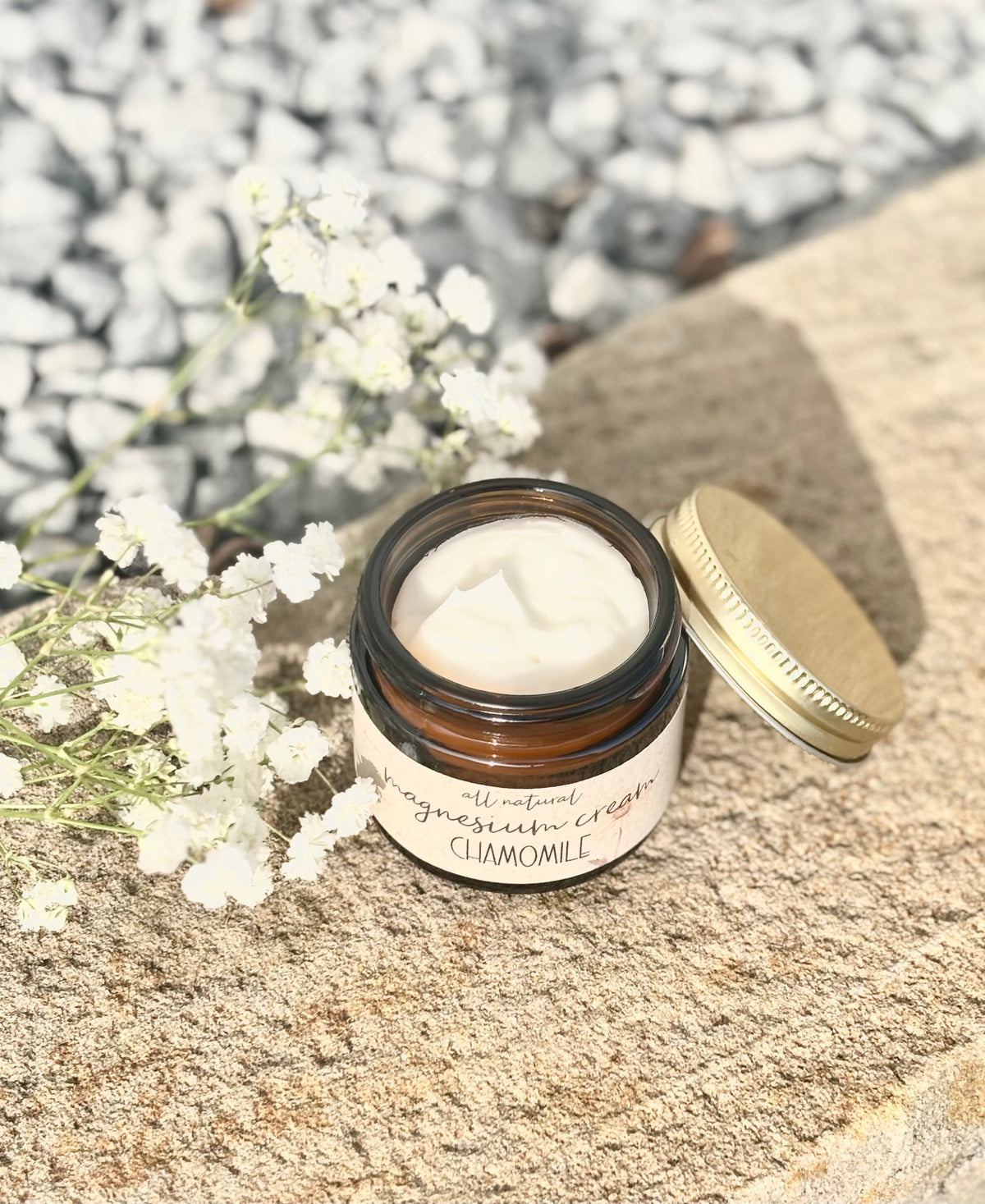 A small amber glass jar of claymole cream with its gold lid resting beside it on a stone surface, surrounded by white baby's breath flowers and a blurred gravel background.