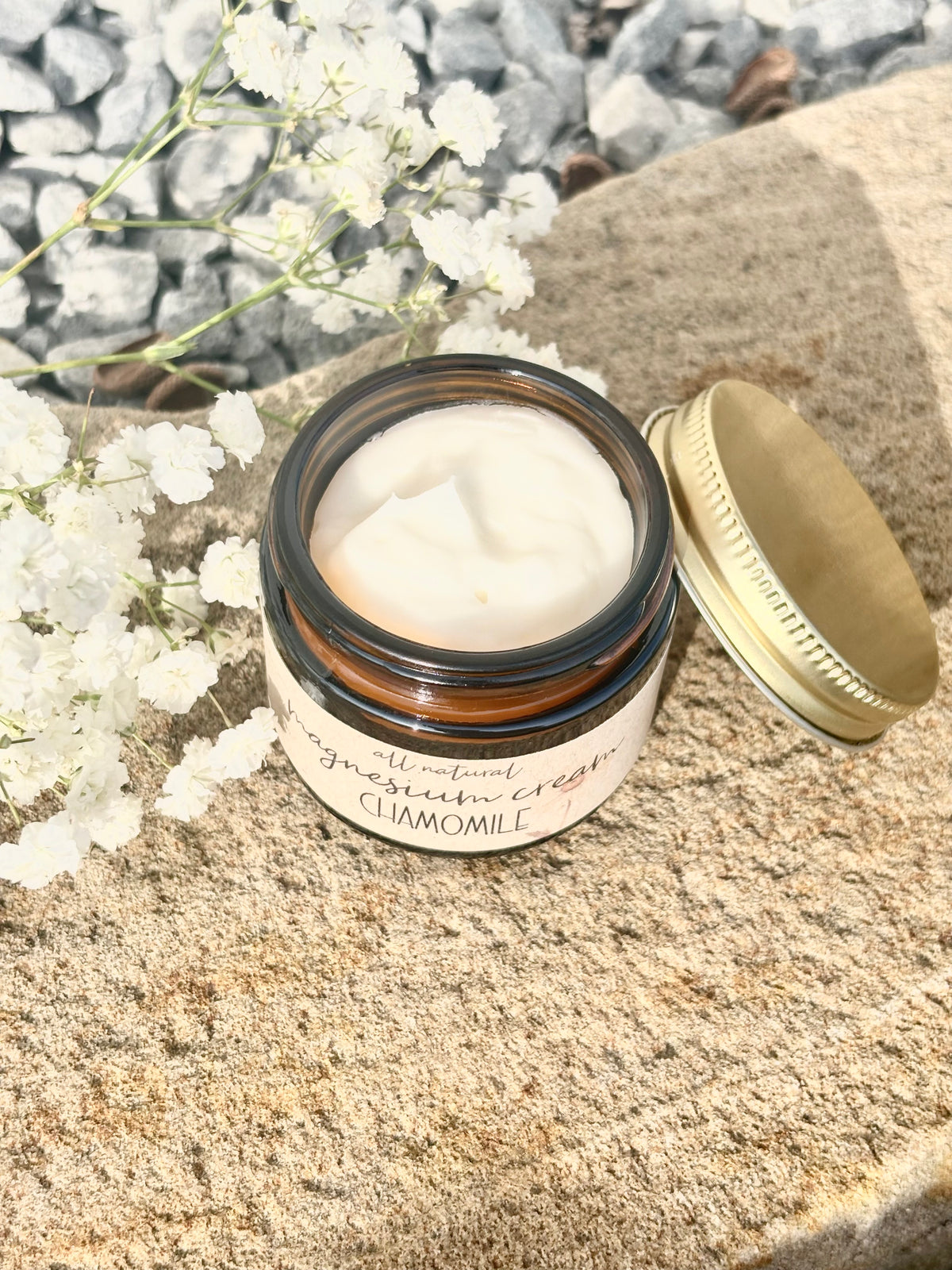 A jar of cream-colored facial mask cream with its gold lid resting beside it on a textured stone surface, decorated with small white flowers.