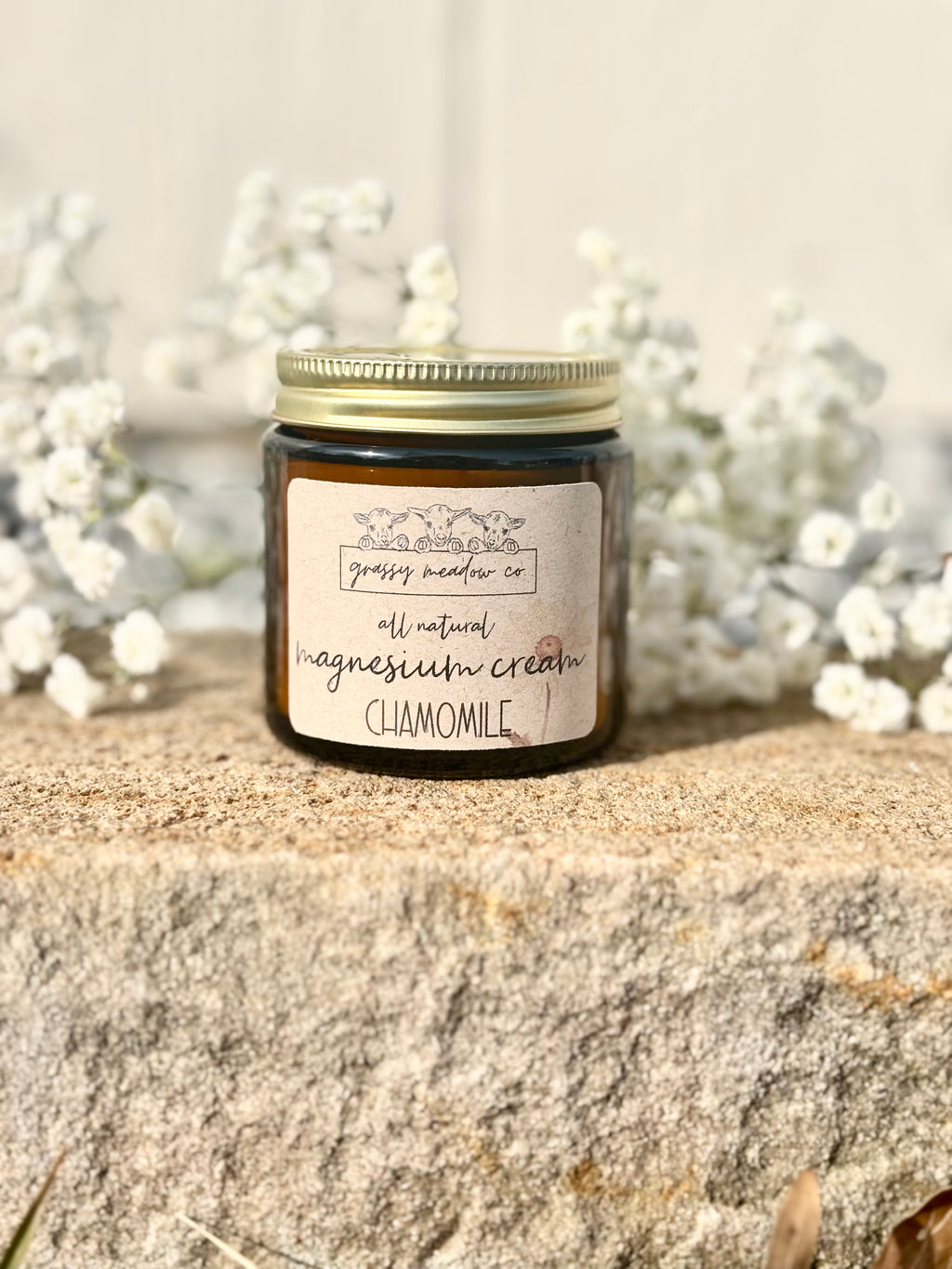 A dark amber glass jar candle with a gold metal lid and a cream label reading 'grassy meadow co all natural magnesium cream CHAMOMILE,' placed on a rough stone surface with white flowers blurred in the background.