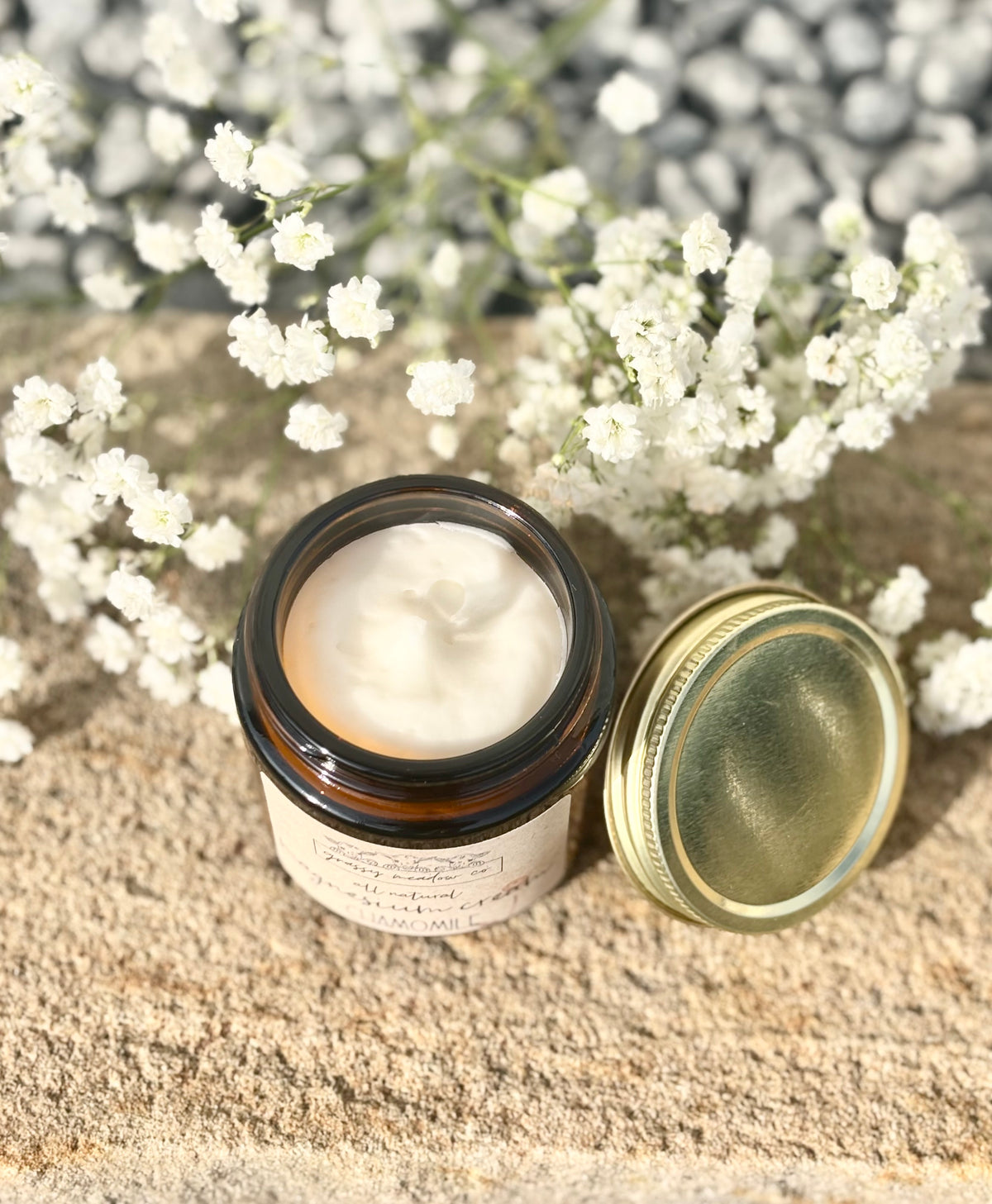 An open amber glass jar candle with a gold lid resting beside it on a textured stone surface, surrounded by small white flowers.