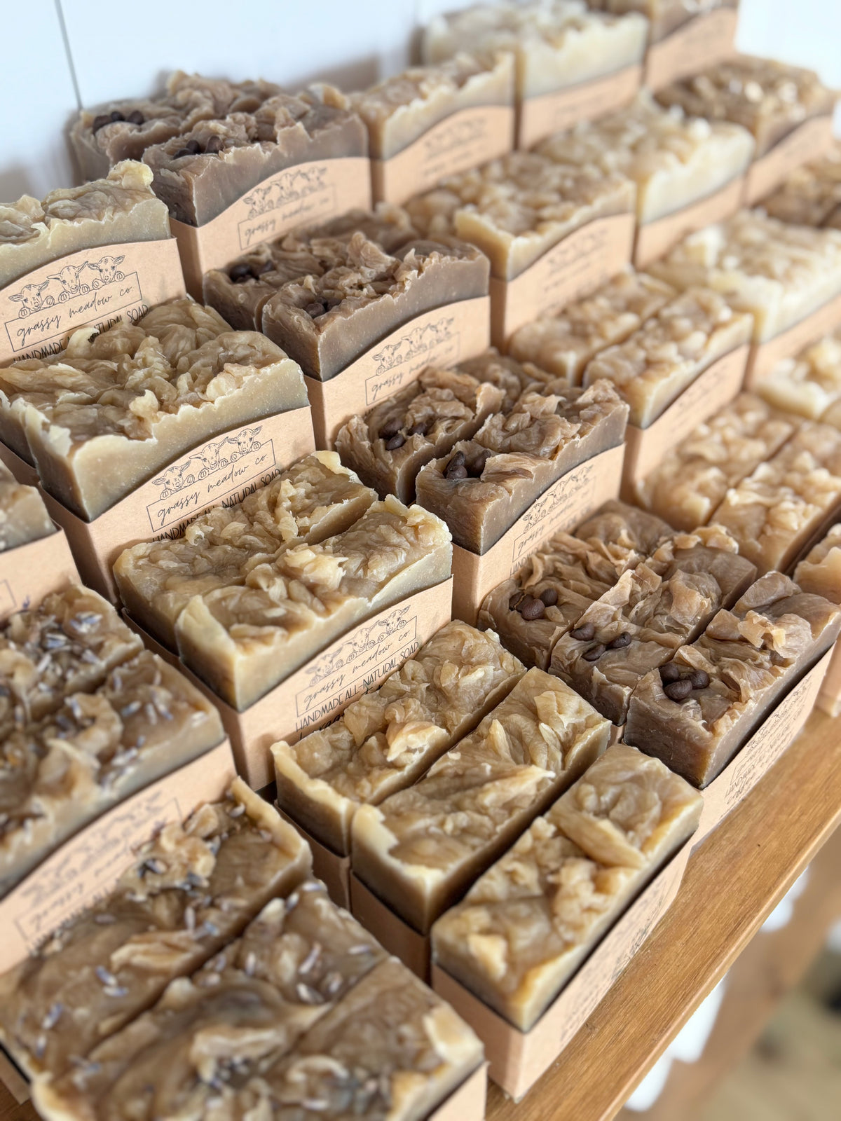 Multiple rectangular bars of handmade soap arranged on a wooden shelf, each wrapped in a kraft paper sleeve with printed text. The soaps have varied earthy tones from beige and tan to brown and gray, with some showing chunky inclusions or layered textures.
