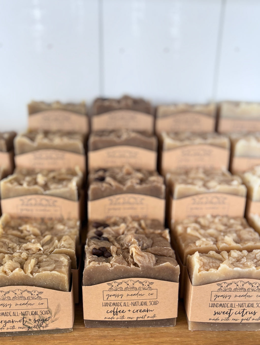 Multiple bars of handmade soap in various shades of brown, beige, and cream, each wrapped in a kraft paper band labeled with the brand 'Grassy Meadow Co.' and scent names like coffee cream, bergamot and sage, and sweet citrus, arranged on a wooden surface.