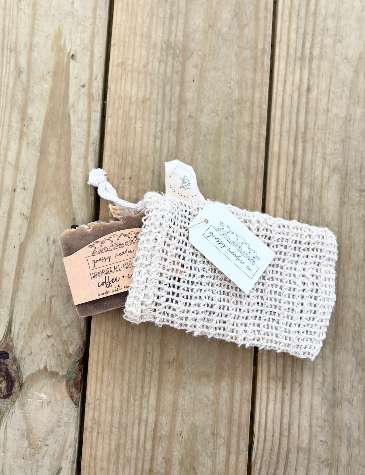 A handmade soap bar wrapped in a cream-colored woven mesh bag, resting on a rustic wooden plank surface. The soap bar is partially visible inside, along with two paper tags attached.