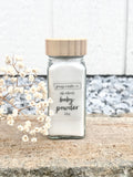 A clear glass jar filled with white baby powder, topped with a light wood lid, placed on a stone surface with dried white flowers and a blurred outdoor background.