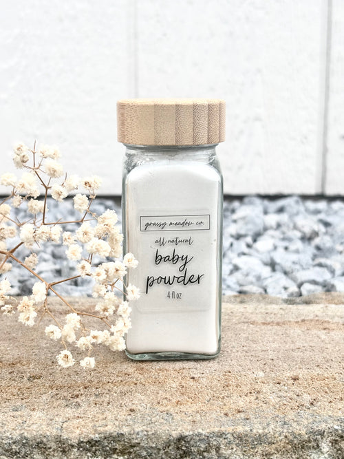 A clear glass jar filled with white baby powder, topped with a light wood lid, placed on a stone surface with dried white flowers and a blurred outdoor background.