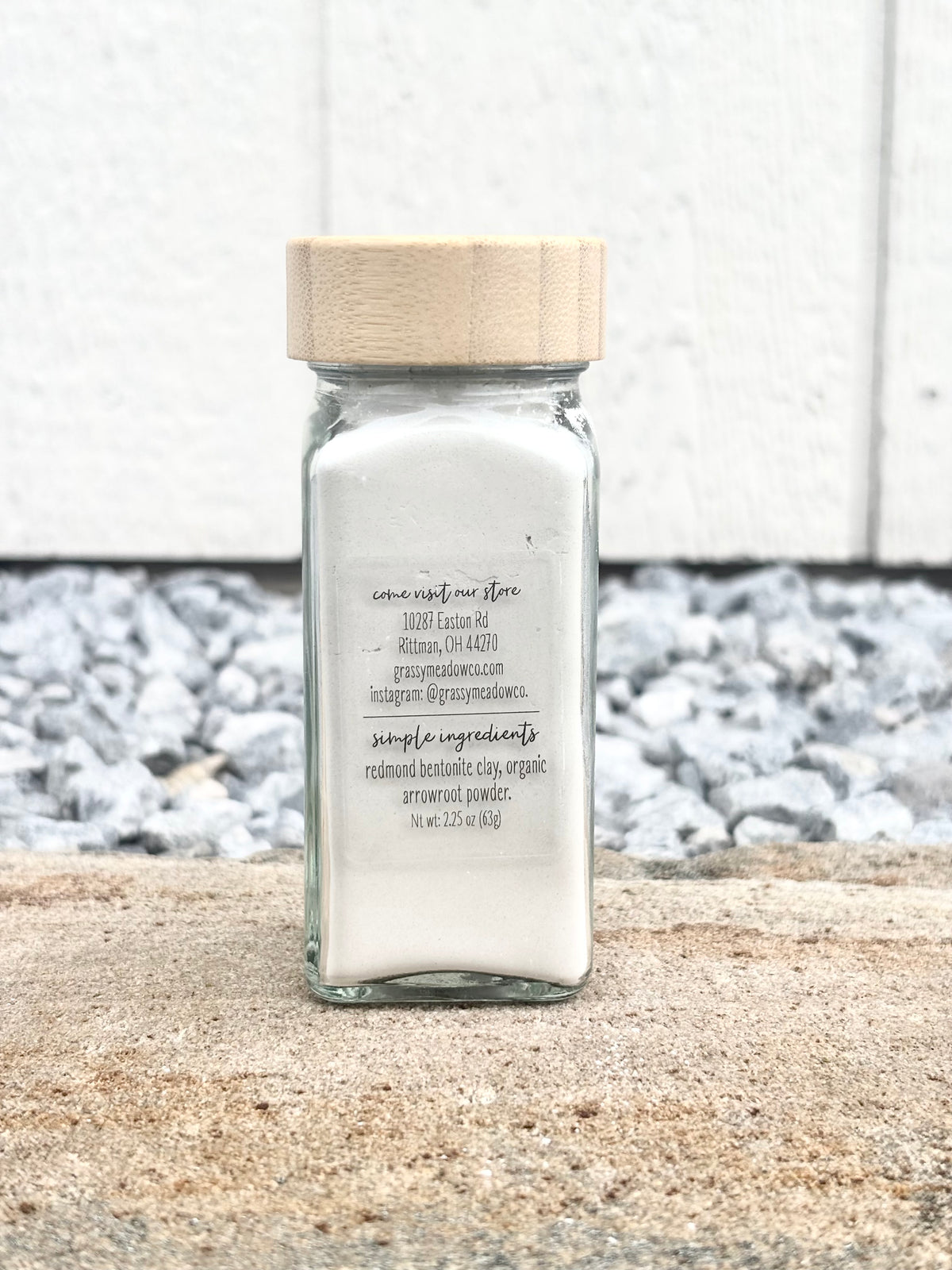 A clear glass jar filled with white baby powder, featuring a light natural wood lid, set on a stone surface against a white textured wall.