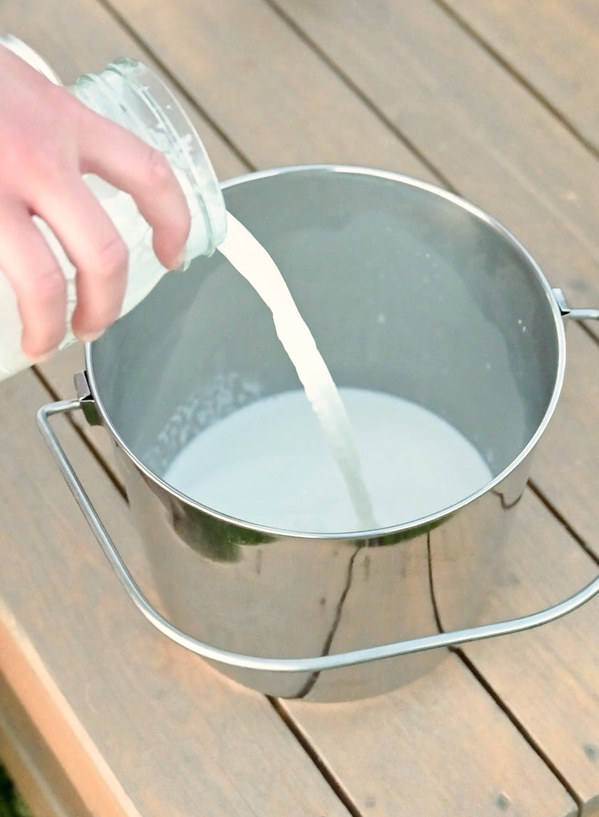 A stainless steel bucket with a wire handle sits on a wooden milk stand surface, with a hand pouring goat milk from a glass bottle into the bucket.