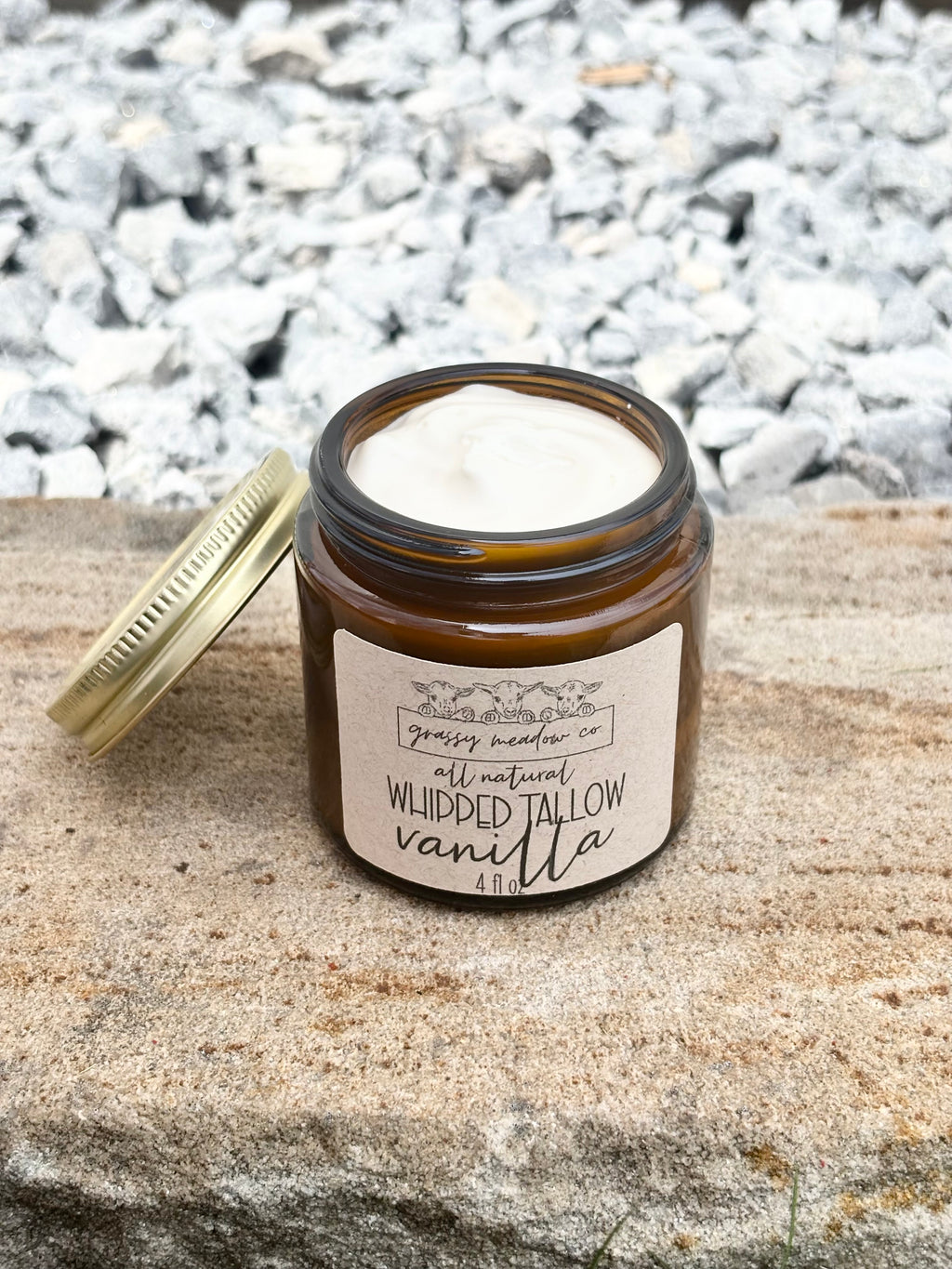 An amber glass jar with a gold lid resting beside it on a stone surface, with a gravel background. The label reads 'Grassy Meadow Co.'