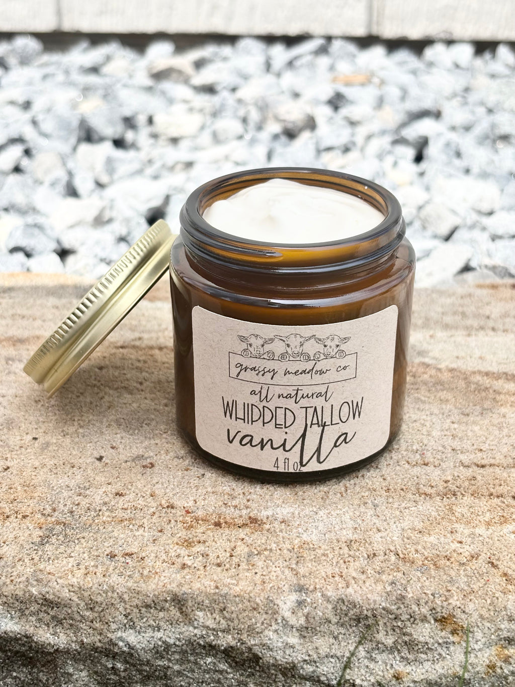 An open amber glass jar with a gold lid leaning beside it on a stone surface outdoors. The label reads Grassy Meadow Co. Whipped Tallow Vanilla