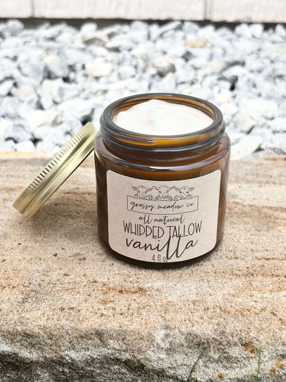 An open amber glass jar with a gold lid leaning beside it on a stone surface outdoors. The label reads Grassy Meadow Co. Whipped Tallow Vanilla