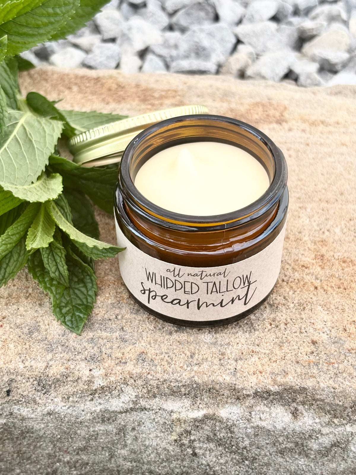 An amber glass jar with a cream-colored lotion and a kraft label reading 'all natural Whipped TALLOW spearmint,' styled on a stone surface with mint leaves and gravel in the background.