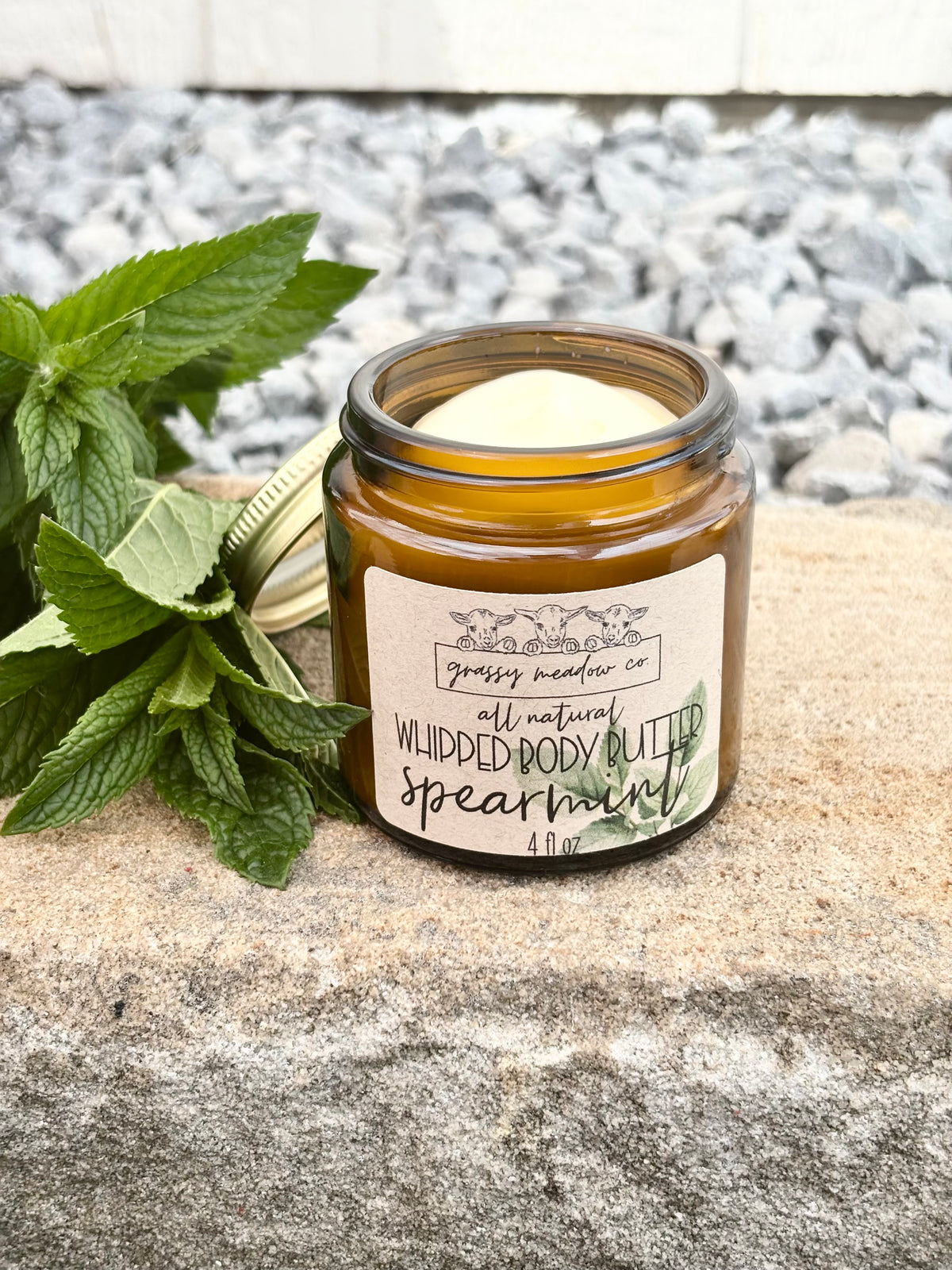 A lotion in an amber glass jar with a cream-colored wax and a white label reading 'Grassy Meadow Co. All Natural Whipped Body Butter, Spearmint.