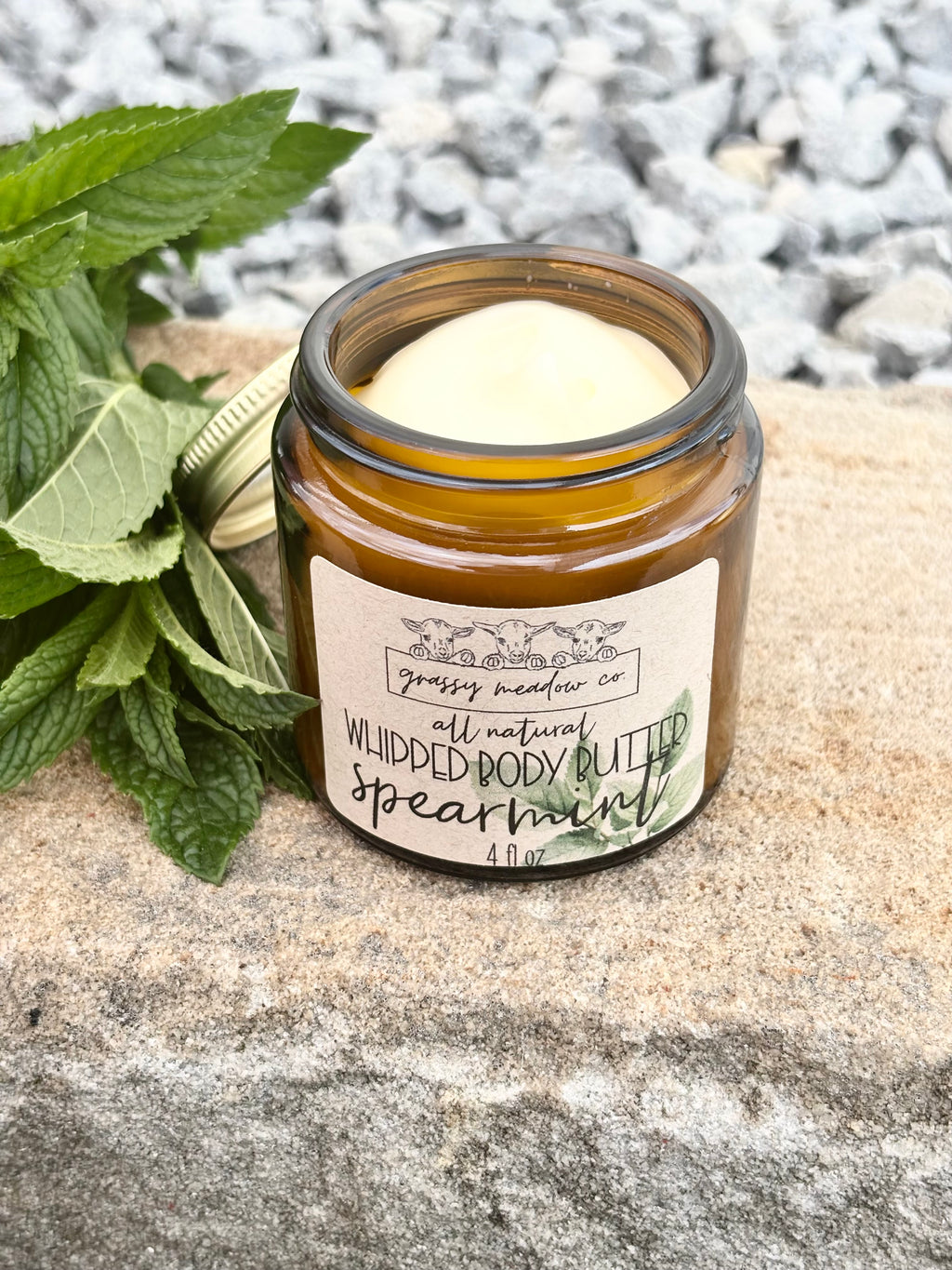 An amber glass jar lotion labeled 'Gassy Meadow Co. All Natural Whipped Body Butter: spearmint' sits on a stone surface next to mint leaves, with gravel in the background.