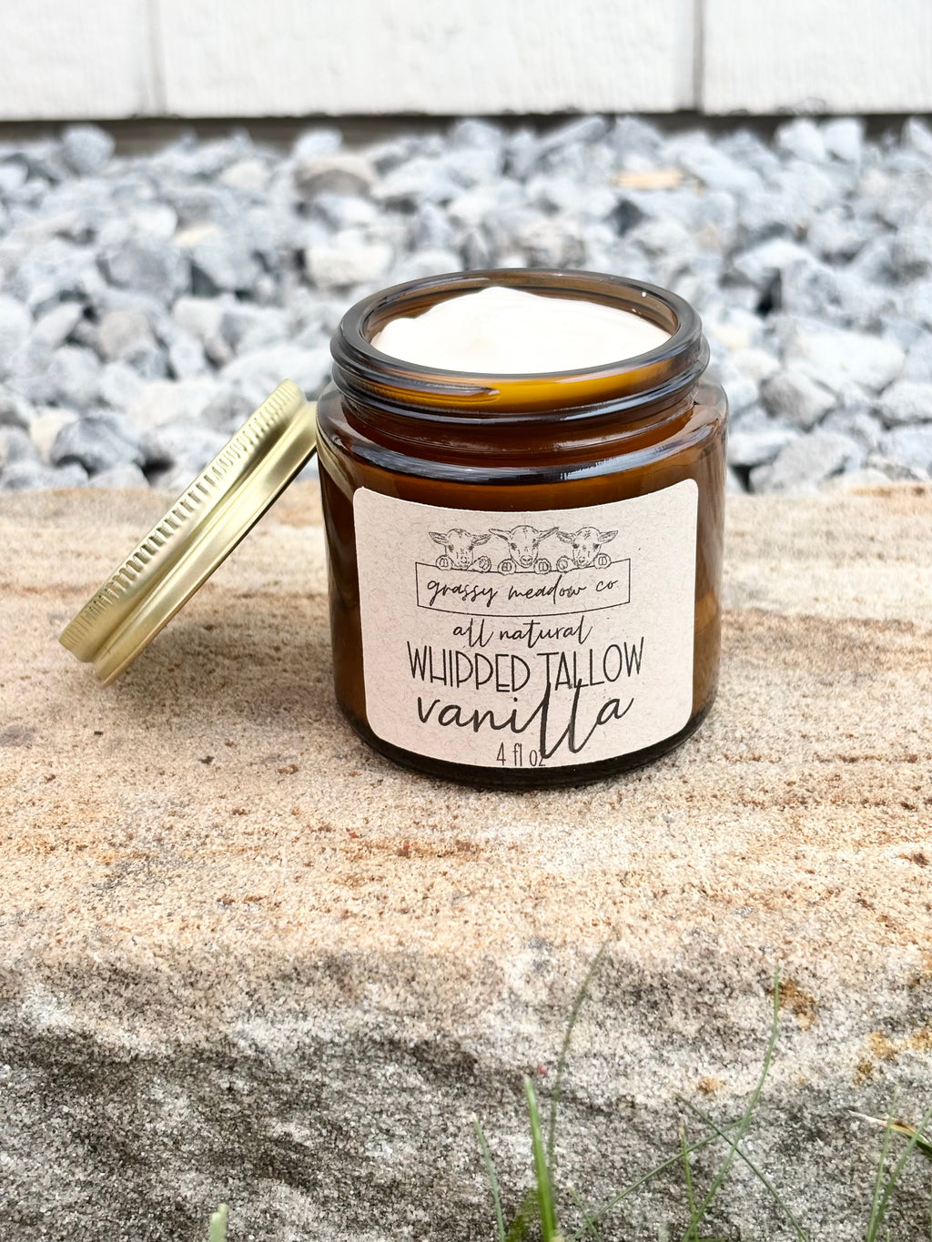 An amber glass jar candle with a gold lid leaning beside it on a stone surface outdoors. The front label reads 'grassy meadow co' and 'all natural WHIPPED TALLOW vanilla'.