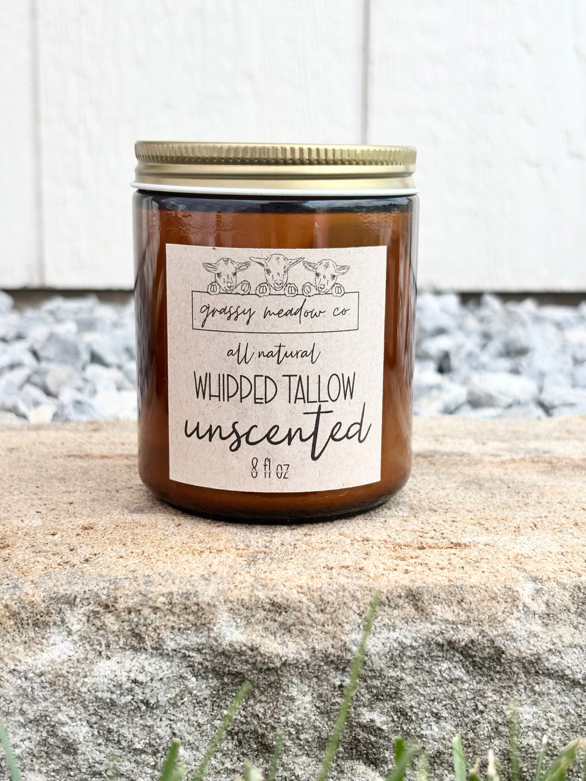 An 8 fl oz amber glass jar filled with whipped lotion with a gold metal lid and a cream paper label reading 'grassy meadow co' and 'all natural WHIPPED TALLOW unscented'. The tallow is placed on a textured stone surface with gravel and a white wooden wall in the background.