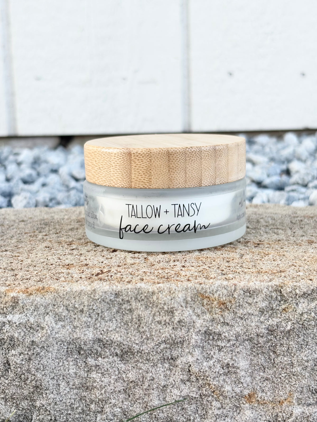 A small jar of Tallow + Tansy face cream sits on a rough stone surface outdoors, with a light wood lid and a clear container showing the white cream inside. The background includes blue-gray gravel and a white paneled wall.