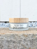 A small jar of Tallow + Tansy face cream sits on a rough stone surface with a white wall and gravel behind it. The jar is frosted with a bamboo lid.