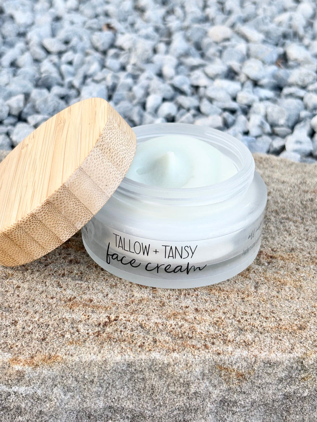 An open jar of Tallow + Tansy face cream sits on a stone surface with a bamboo lid leaning beside it, and blurred gray pebbles in the background.