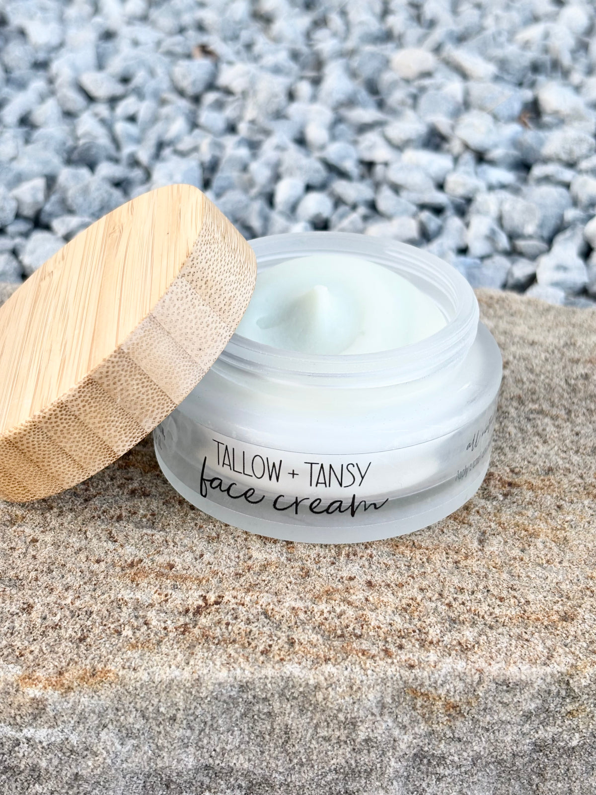 An open jar of Tallow + Tansy face cream sits on a stone surface with a bamboo lid leaning beside it, and blurred gray pebbles in the background.